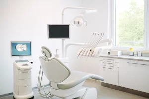 Contemporary dental clinic with a central dental chair, two monitors, overhead light, and organized cabinetry. Natural light enters through a large window, highlighting the clean and professional setup.