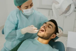 Close-up view of a dental patient lying on a treatment chair with eyes closed while a dentist, wearing blue gloves and a white coat, uses a dental scaler and suction tool to clean the patient's teeth. The focus is on the patient’s open mouth and the dentist’s hands performing the procedure.